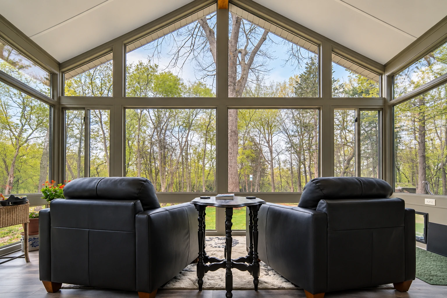Bright four-season sunroom with tall angled windows, two black recliners, and a view of leafy trees.
