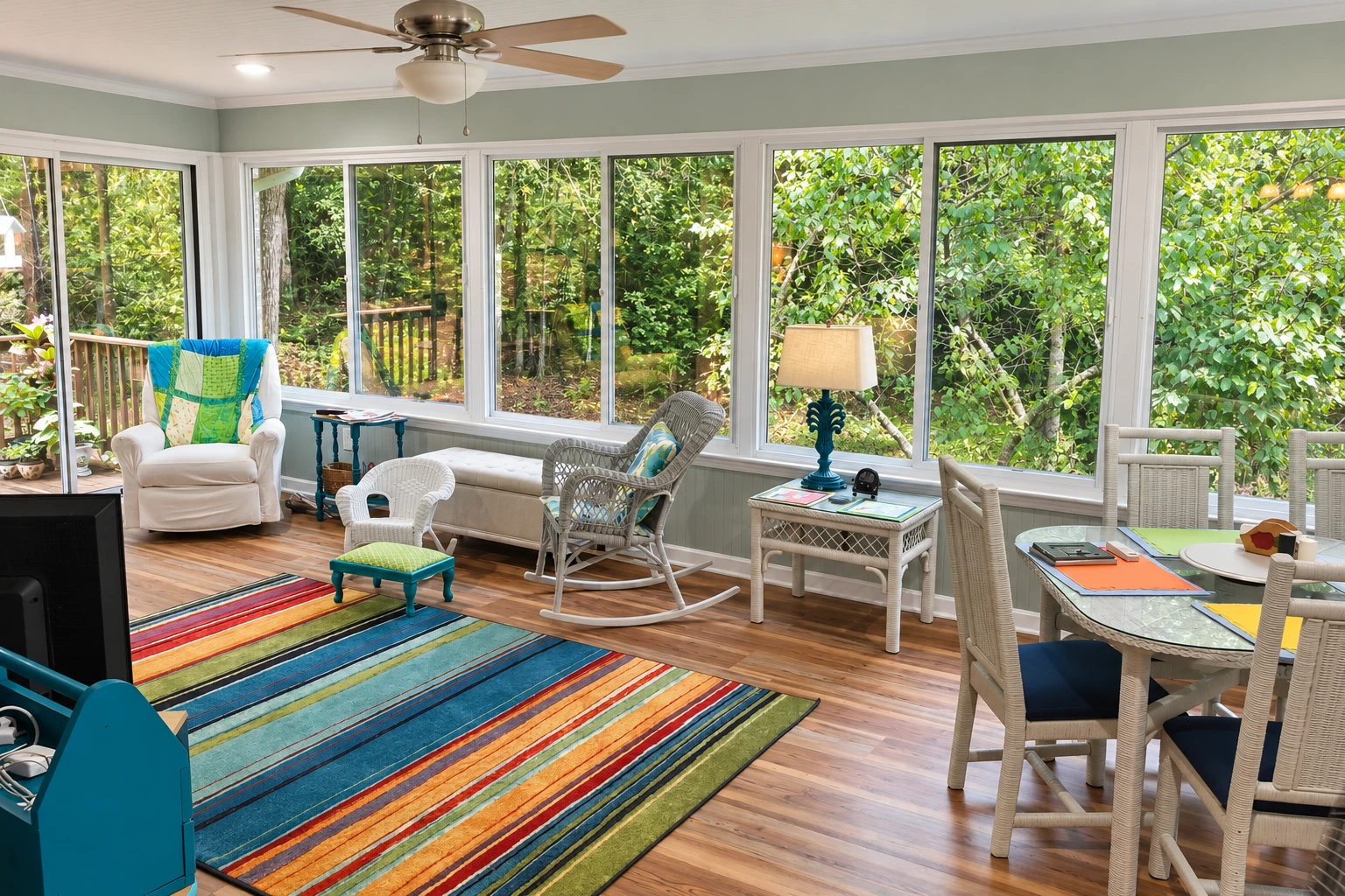 A furnished sunroom with broad windows overlooking dense green trees, a striped rug, and wicker seating.