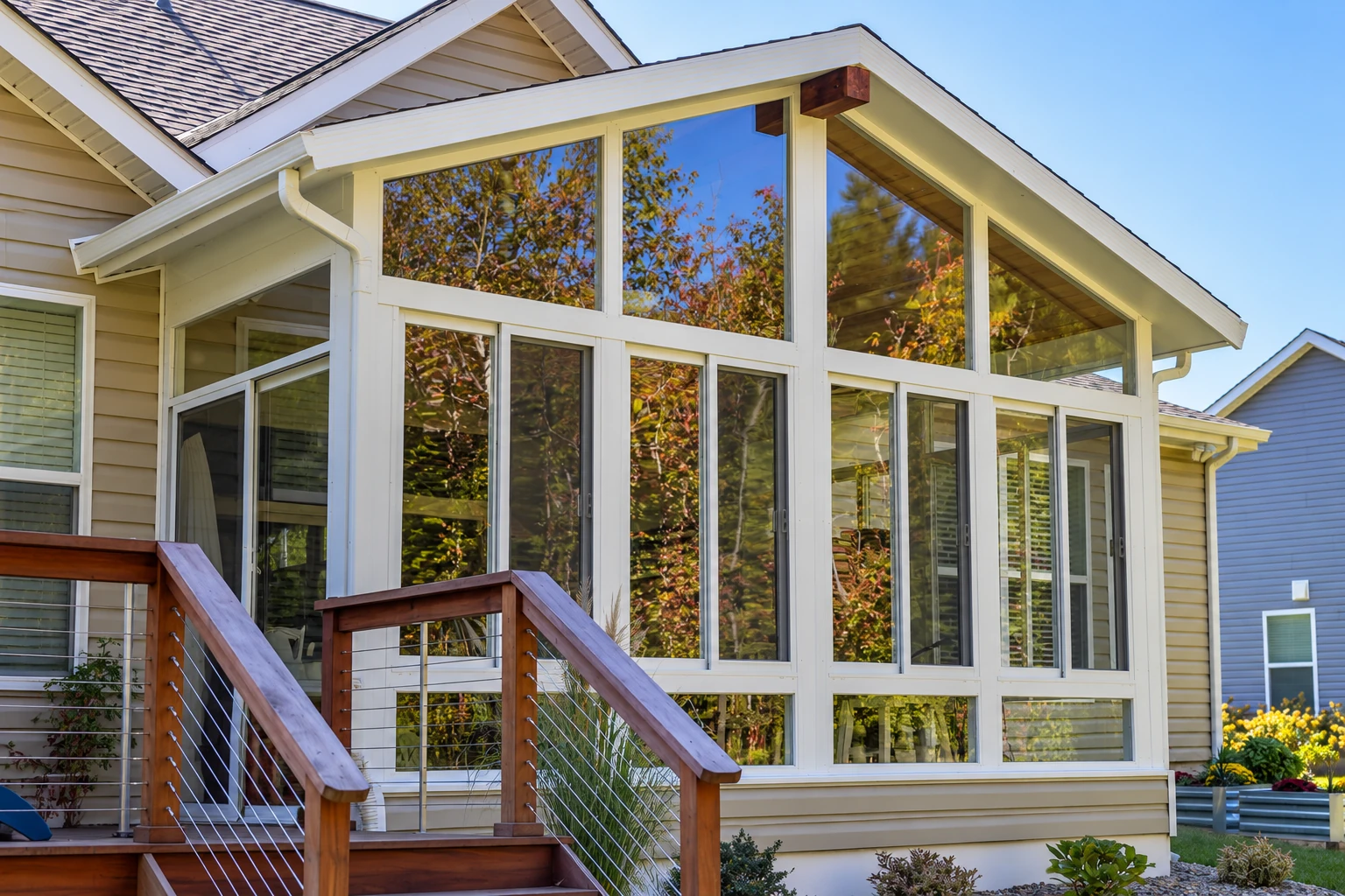 White-framed, gable-roofed four-season sunroom with tall windows and wooden deck steps on a beige-sided house.