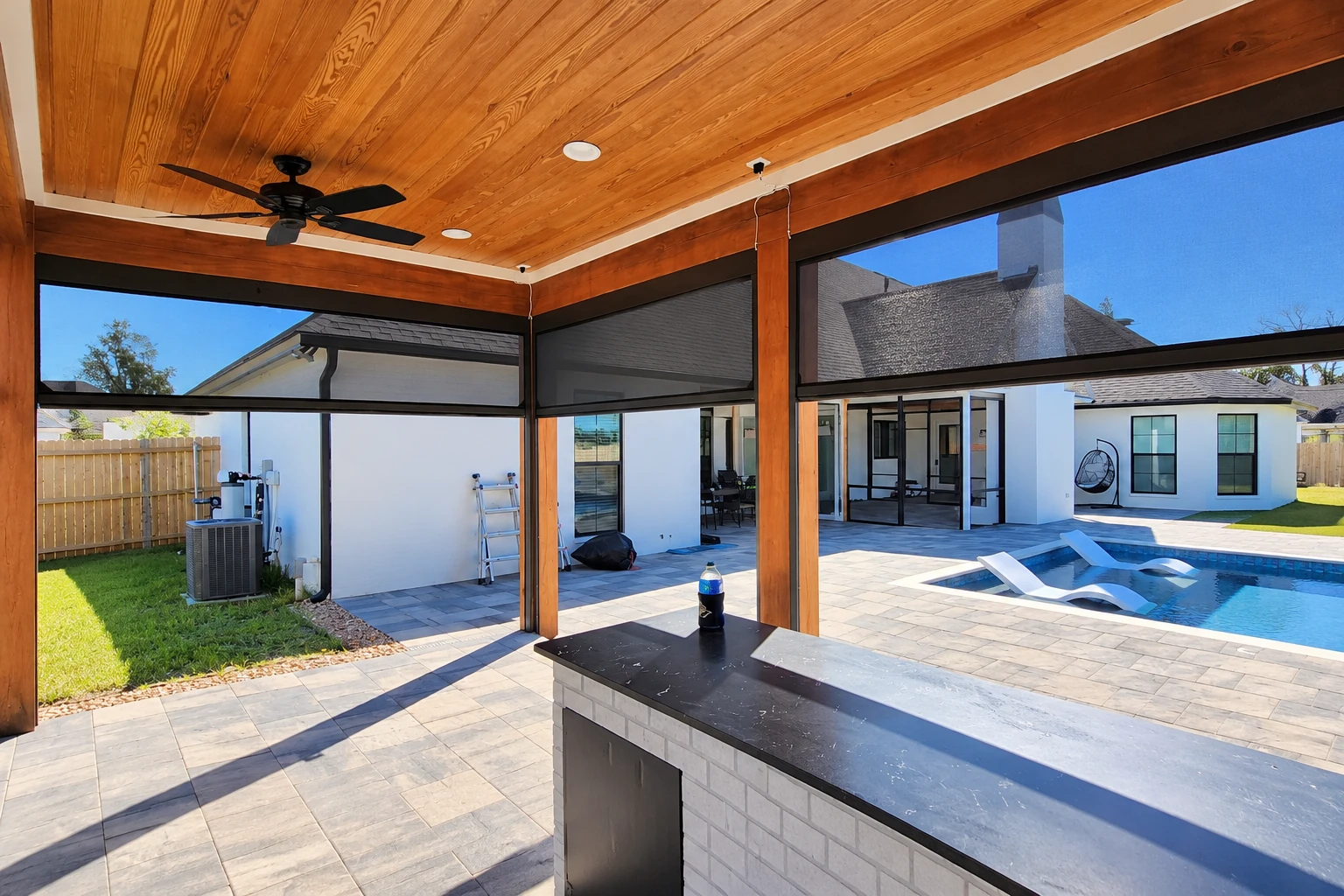 Covered patio with a wood-paneled ceiling, ceiling fan, retractable screens, and a poolside kitchen counter.