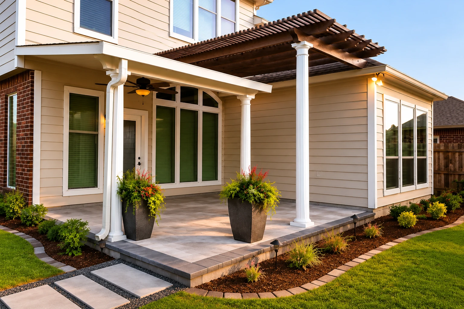 A tiled patio beneath a dark wooden pergola and white roof extension is furnished with planters beside a beige-sided house.