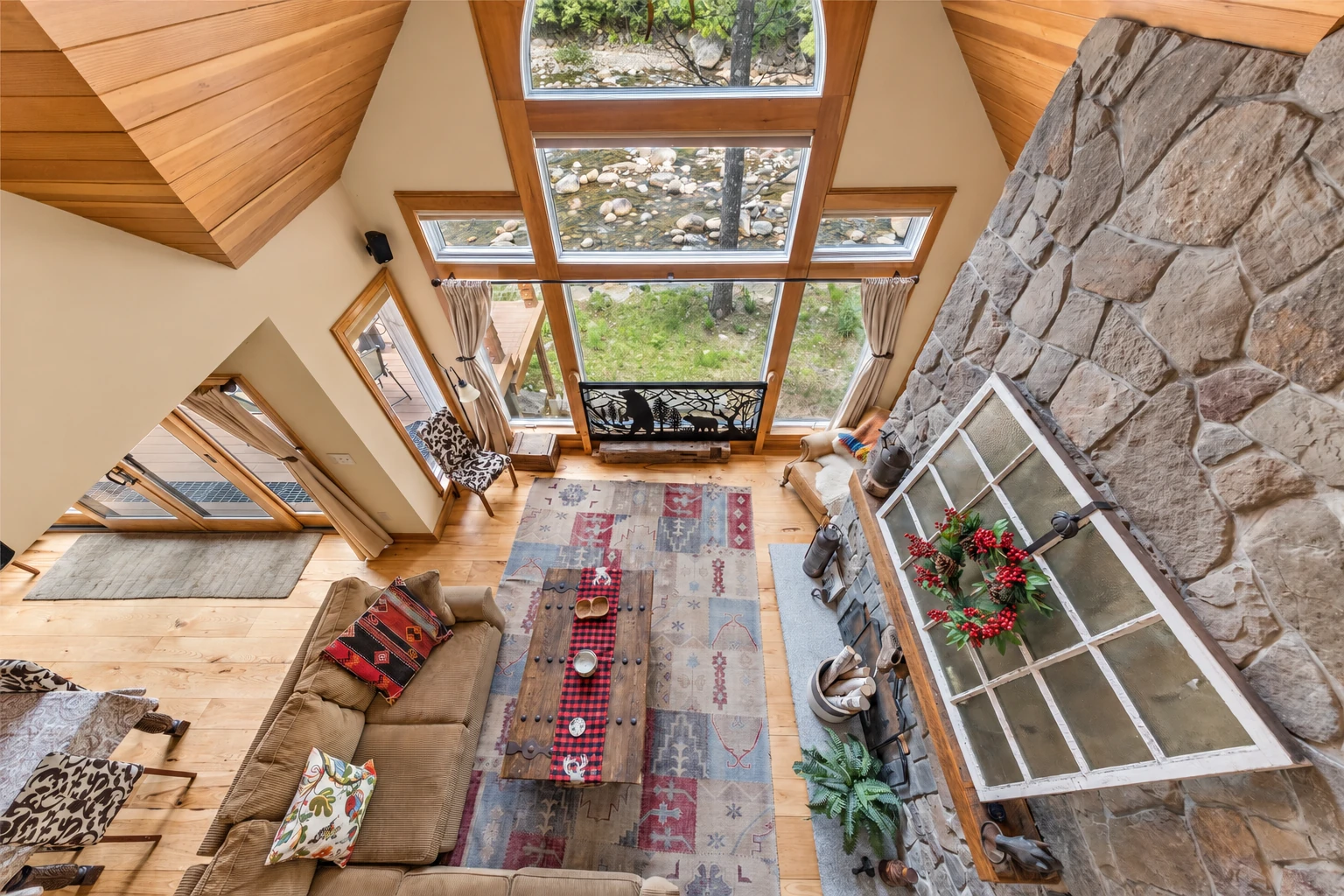 A vaulted sunroom interior has a tall wall of windows, warm wood ceilings, a stone fireplace, and seating around a rug and coffee table.