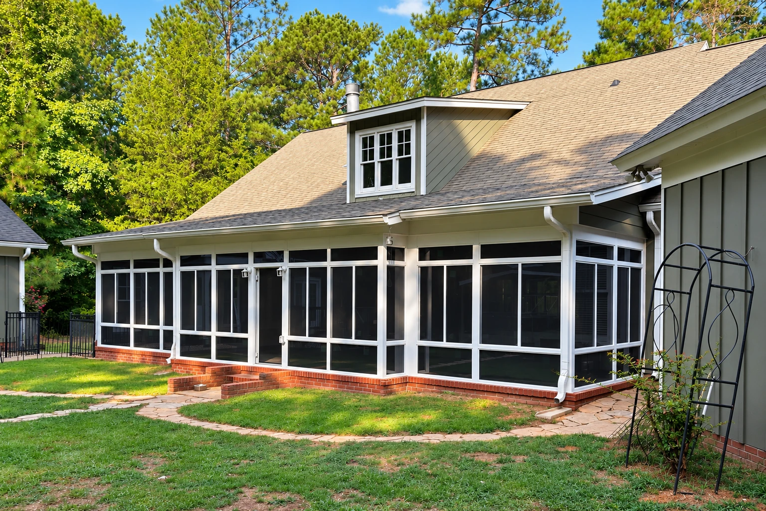 A white-framed sunroom addition with dark screened windows extends from a shingled house into a grassy, tree-lined yard.