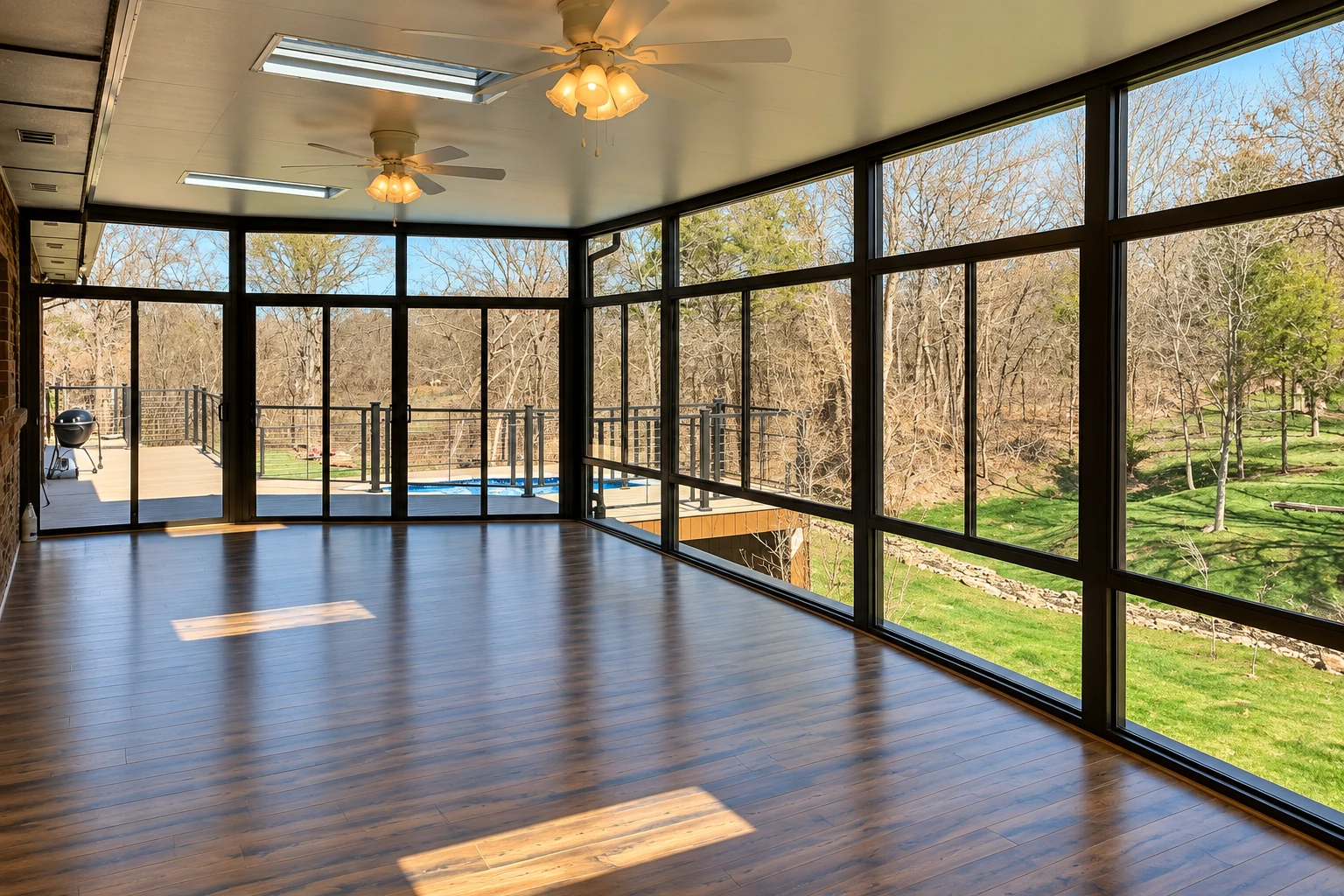 An empty sunroom with dark-framed floor-to-ceiling windows, wood floors, and views of a yard and pool.